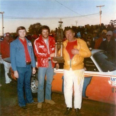 Both Robert Gee and Dale Earnhardt Sr. are holding the trophy in front of their car in this old picture.